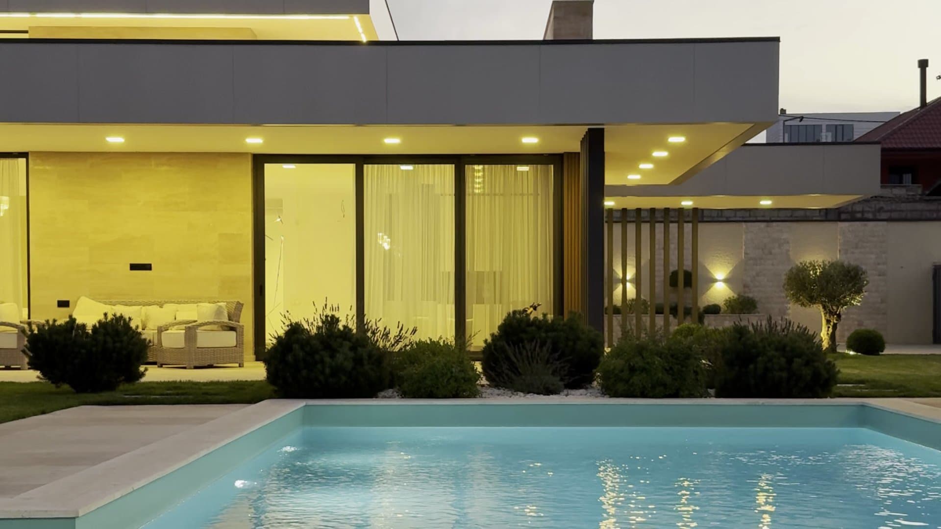 The house at dusk, its glazed rooms lit, beyond the pool